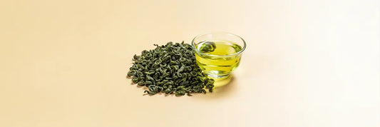 A cup of brewed tamaryokucha green tea with pale yellow-green liquor beside a small pile of curled comma-shaped dry leaves, representing the low-astringency Japanese green tea known for its calm energy and antioxidant benefits.