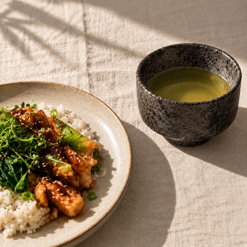 A close-up of a plated meal with rice, glazed chicken, and green vegetables beside a speckled dark ceramic cup of light green tea on a beige linen tablecloth.