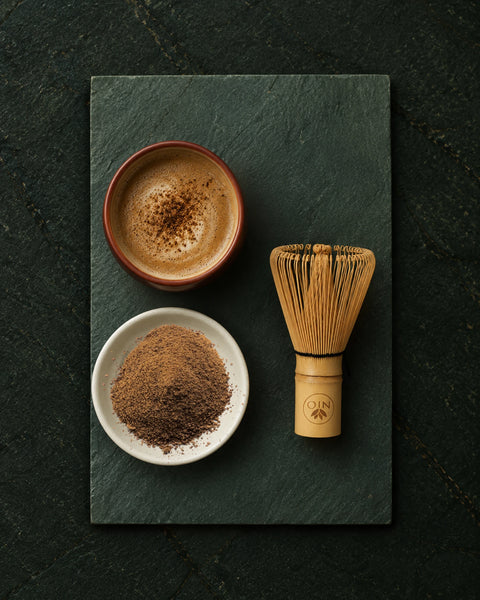 A bamboo whisk and a small bowl of whisked hojicha powder are arranged on a dark slate board next to a cup of hojicha latte with foam, on a dark textured background.
